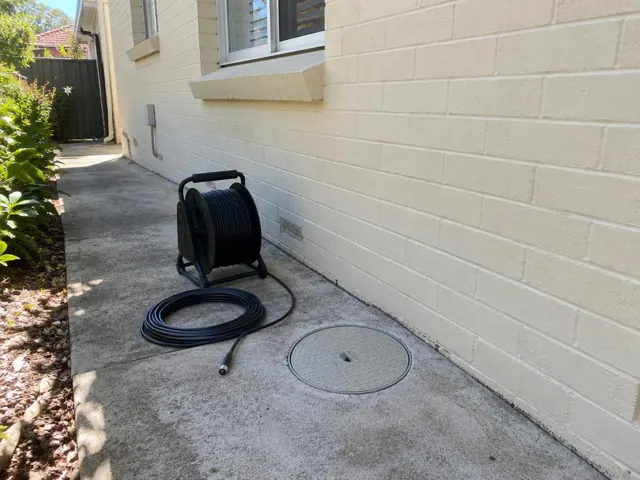 Capped drain inspection opening beside a coiled camera reel along a brick house wall in Thornleigh