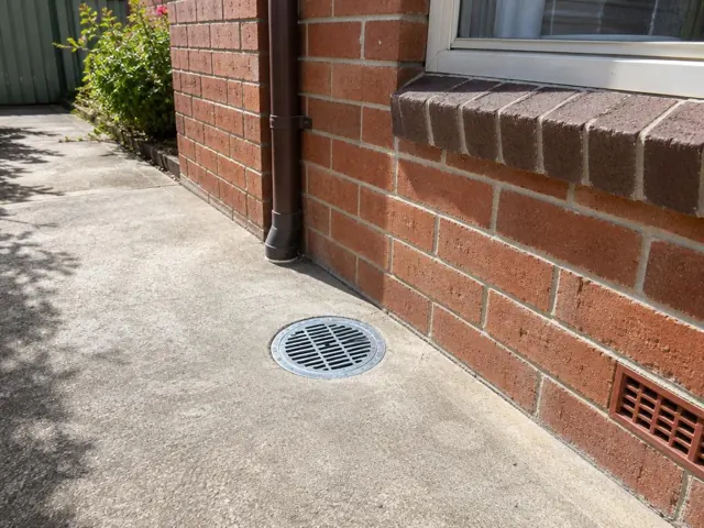 Outdoor gully grate beside a brick house wall and concrete path in Normanhurst