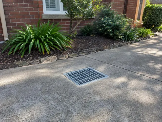 Concrete driveway with a seated stormwater grate beside a brick house and garden bed in Hornsby