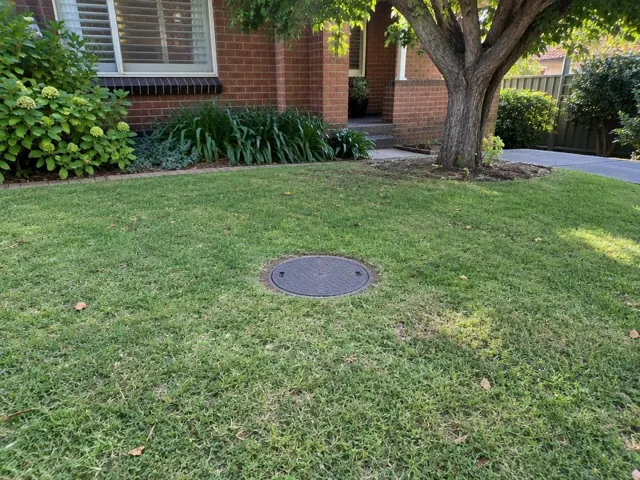Circular sewer access cover set in a tidy grass lawn beside a brick house in Thornleigh